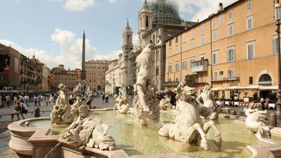 Fontana monumentale con statue in marmo in una grande piazza urbana, con acqua in vasca e sculture di figure mitologiche. Sullo sfondo si vedono edifici storici e una chiesa con cupola e impalcature, oltre a un obelisco al centro della piazza.