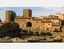 Borgo medievale in pietra su una collina, con edifici dai tetti in tegole e una grande porta ad arco nelle mura. Sullo sfondo si vedono torri quadrate e case addossate, con alberi e vegetazione ai piedi delle fortificazioni.