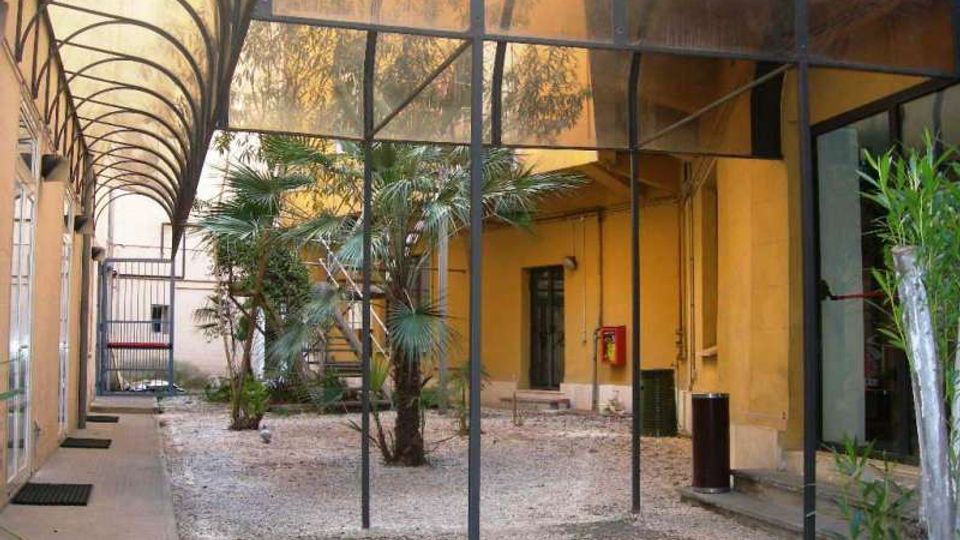 Cortile interno di un edificio con pavimento in ghiaia e piccole palme al centro, circondato da muri gialli con porte e finestre. Una tettoia arcuata in metallo e pannelli traslucidi copre il passaggio laterale e parte dello spazio, con pali verticali e un cancello sul fondo.