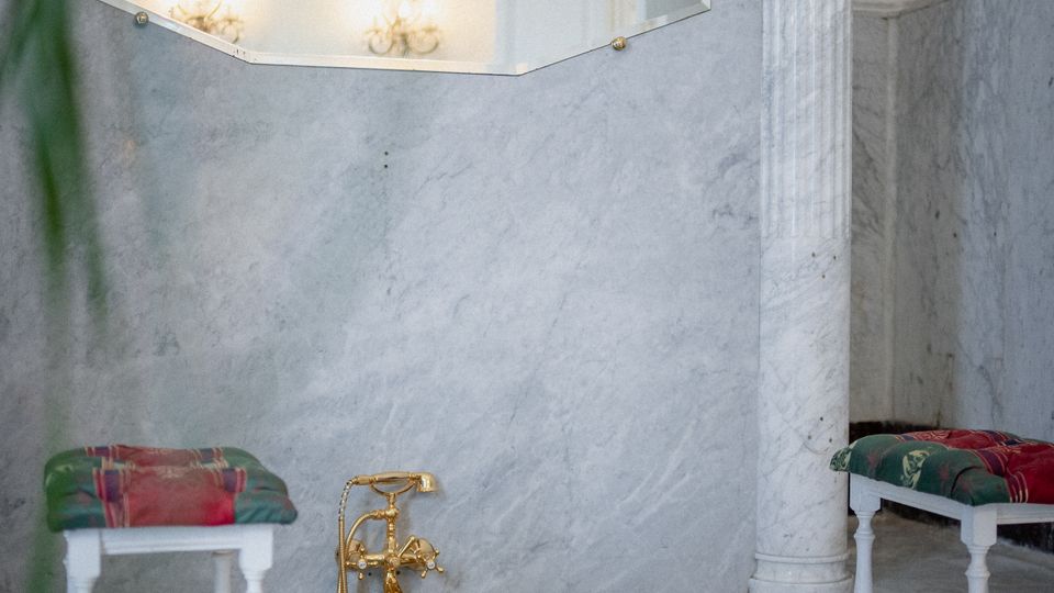 Interno di un bagno in marmo con vasca incassata e rubinetteria dorata, affiancata da due piccoli sgabelli con cuscini. Sulla parete c’è uno specchio esagonale con luci riflesse e una colonna in marmo vicino al bordo della vasca.