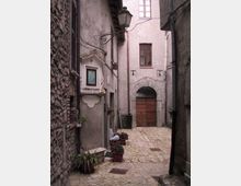 Vicolo stretto in un borgo storico con pavimentazione in pietra, muri in muratura grezza e intonaco, e cavi a vista. In fondo si vede un grande portone ad arco in pietra con porta in legno, una lanterna sporgente e vasi di piante lungo le pareti.