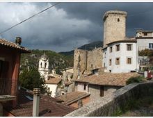 Veduta di un borgo in pietra con tetti in coppi, una torre cilindrica medievale e resti di mura, affiancati da edifici intonacati con finestre. Sullo sfondo si vedono colline boscose e un cielo nuvoloso.