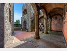 Cortile in pietra di un edificio storico con portico ad archi sostenuto da colonne e soffitto a volte in mattoni. Sul lato destro ci sono botti accatastate e una parete in mattoni, mentre a sinistra si vedono mura in pietra, un cancello ad arco e una balaustra con vasi.