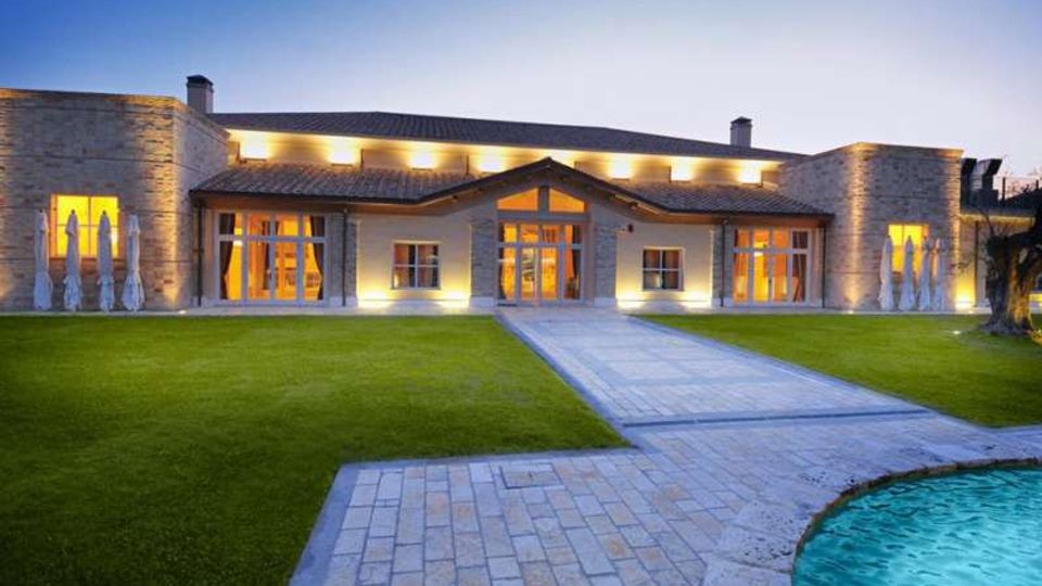 Edificio moderno a un piano con rivestimento in pietra e ampie vetrate illuminate, affacciato su un prato curato con vialetto in pietra che conduce all’ingresso centrale. In primo piano si vede il bordo di una piscina e, sullo sfondo, il cielo al crepuscolo.