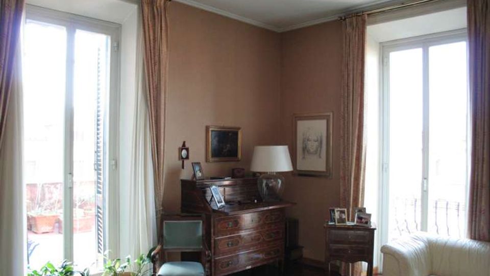 Interno di un soggiorno con due grandi portefinestre affacciate su un balcone, tende lunghe e pareti color tortora. Al centro ci sono un mobile in legno tipo secretaire con lampada e quadri appesi, una sedia, un piccolo tavolino con cornici e una poltrona chiara; alcune piante sono vicino alla finestra.