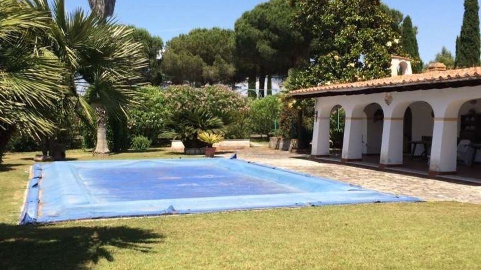 Giardino di una villa con prato e piscina rettangolare coperta da un telo azzurro, circondata da palme e alberi. A destra si vede un portico bianco con archi e tetto in coppi, con vialetto in pietra davanti.