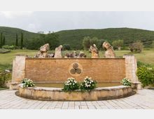 Fontana semicircolare in pietra con vasca bassa e parete in mattoni decorata da un emblema circolare; sopra la parete sporgono grandi rocce ornamentali. Davanti sono disposte fioriere con fiori bianchi e sullo sfondo si vedono colline verdi e alberi in un paesaggio rurale.