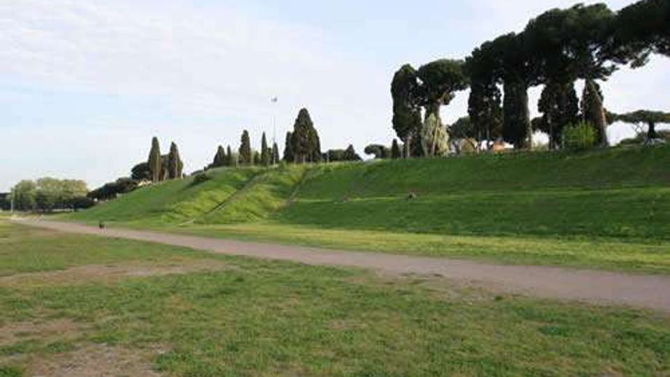 Ampio parco con prato in pendenza e un sentiero sterrato che attraversa l’area. Sullo sfondo si vedono alberi alti e sagomati, tra cui cipressi e pini, lungo il crinale erboso.