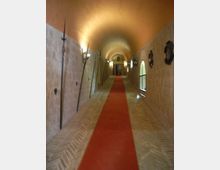 Lungo corridoio in pietra con volta a botte e pavimento in mattoni, attraversato da un tappeto rosso centrale che conduce a una porta in fondo. Sulle pareti sono appese armi decorative e lampade, con alcune finestre ad arco sul lato destro.