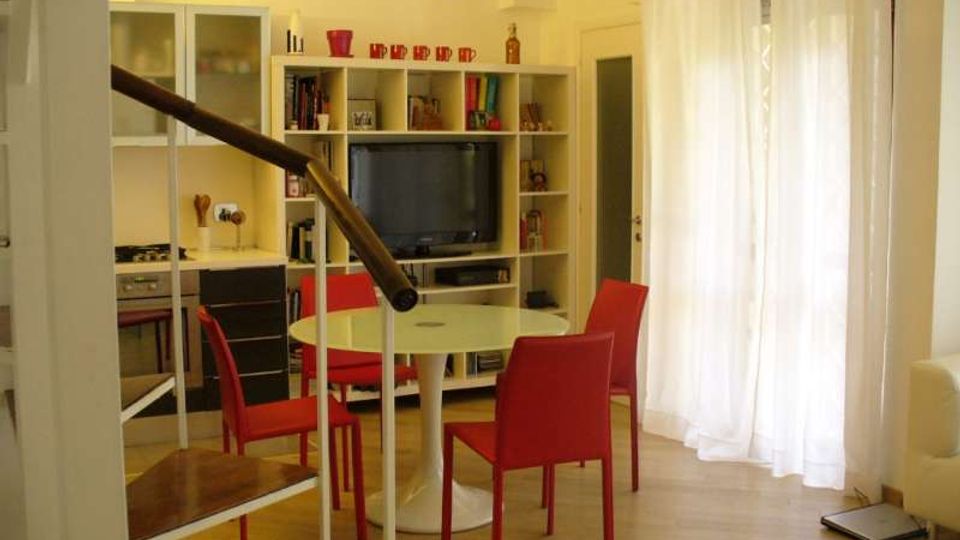 Soggiorno moderno open space con angolo cottura a sinistra, tavolo rotondo bianco con sedie rosse e libreria a parete con TV. A destra una grande portafinestra con tende bianche illumina il pavimento chiaro; in primo piano si vede la ringhiera di una scala interna.