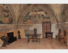 Interno di una sala storica con soffitto a volta affrescato e lunette dipinte, pareti decorate e pavimento in cotto. Al centro ci sono tavoli e sedie in legno, con una porta a pannelli sul fondo e arredi scuri lungo le pareti.