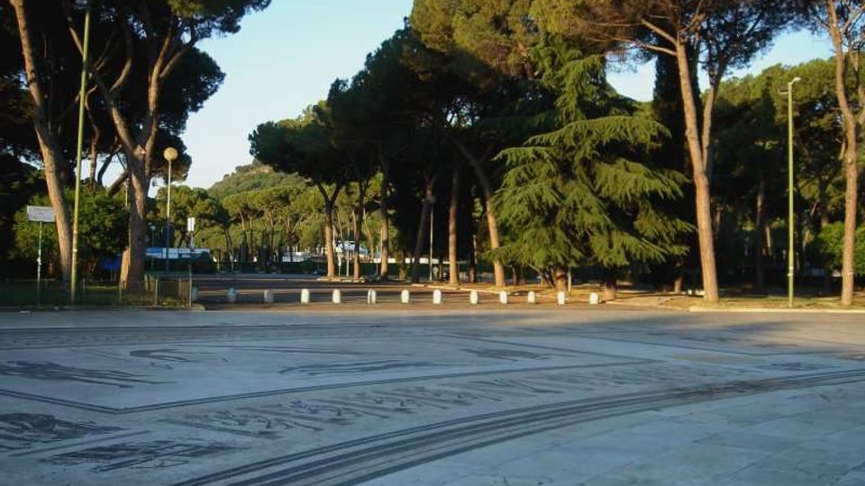 Ampia piazza pavimentata in pietra con motivi geometrici scuri, circondata da alberi ad alto fusto e da un filare di pini. Sullo sfondo si vedono lampioni, una strada e una fila di dissuasori bianchi lungo il bordo della piazza.