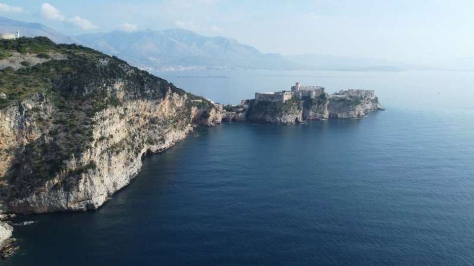 Veduta costiera con alte scogliere rocciose che scendono nel mare calmo e, al largo, una fortezza in pietra su un isolotto collegato alla terraferma. Sullo sfondo si intravedono montagne e una costa lontana sotto un cielo limpido.