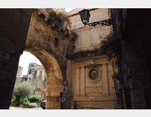 Antico passaggio ad arco in pietra che collega edifici in muratura, con una facciata decorata da un’edicola con medaglione incassato e cornici scolpite. In alto si vede un lampione in ferro battuto e, sullo sfondo, una piccola piazza con vegetazione e costruzioni storiche.