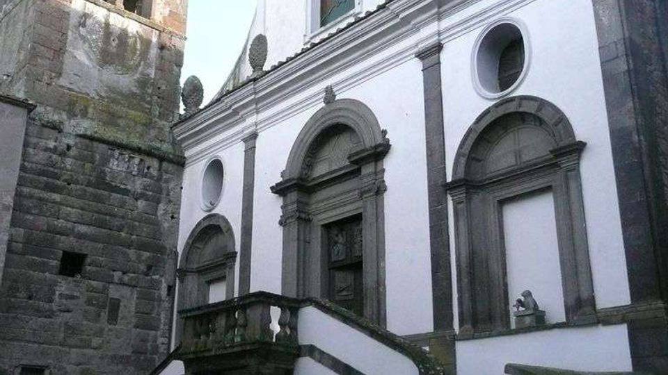 Facciata di una chiesa in stile barocco con intonaco bianco e cornici in pietra scura, ingresso sopraelevato raggiunto da una scalinata doppia curva con balaustra. A sinistra si vede un campanile in pietra con finestre ad arco, in una piccola piazza pavimentata.