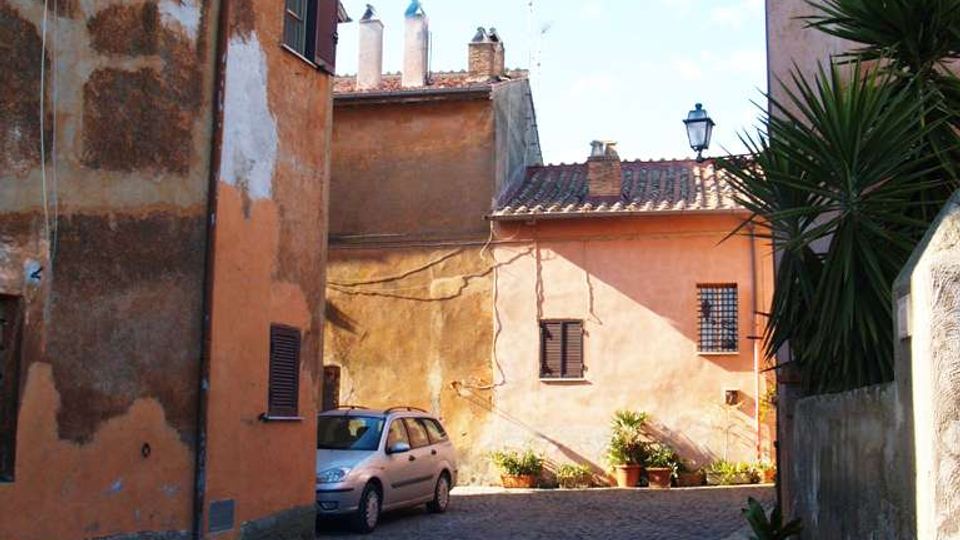 Vicolo stretto in pietra con pavimentazione in sampietrini, fiancheggiato da edifici intonacati color ocra e rosa con persiane scure e tetti in coppi. In fondo si vedono alcune piante in vaso, un lampione e una piccola auto parcheggiata lungo il muro.