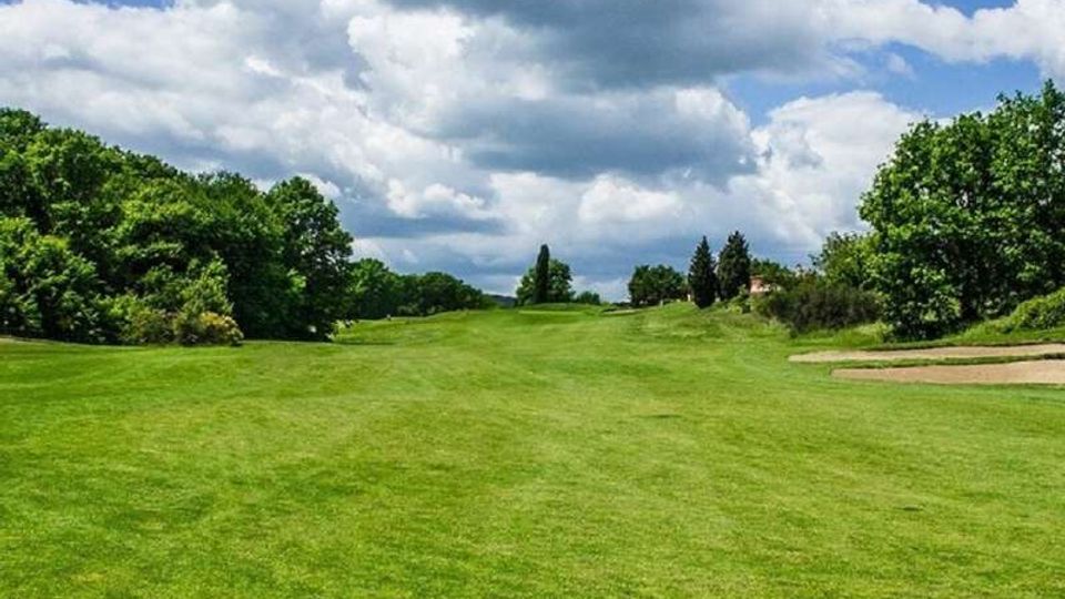Ampio campo da golf con fairway erboso in leggera salita, circondato da alberi e cespugli ai lati. A destra si vede un bunker di sabbia, con cielo azzurro e grandi nuvole sullo sfondo.