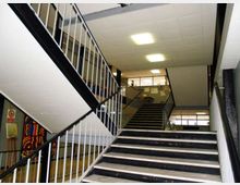 Interno di un edificio scolastico con ampia tromba delle scale: gradini scuri con bordi chiari e rampe sovrapposte con corrimano e ringhiere metalliche bianche e nere. Soffitto a pannelli con luci rettangolari e grandi finestre sul pianerottolo superiore.