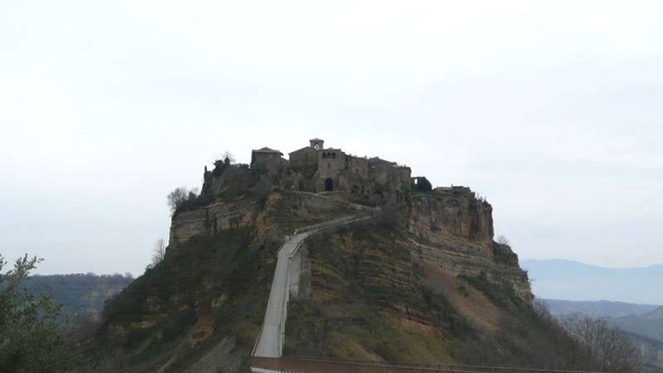Borgo e complesso di edifici in pietra su una rupe rocciosa, collegati da una lunga strada sopraelevata che sale fino all’ingresso. Sullo sfondo si vedono colline e una valle sotto un cielo coperto.