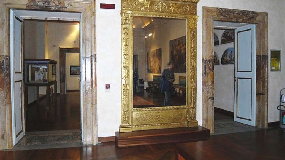 Interno di museo con pavimento in parquet scuro e pareti bianche, con due porte aperte incorniciate in marmo che conducono a sale espositive. Al centro è presente un grande specchio con cornice dorata decorata, su pedana in legno, e sulle pareti si intravedono dipinti e teche.
