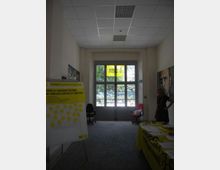 Atrio interno con soffitto a pannelli e una grande porta a vetri che affaccia su alberi all’esterno, con uno striscione giallo “Amnesty International” sopra l’ingresso. Ai lati si vedono un pannello informativo e un tavolo con materiali e volantini su tovaglia gialla.