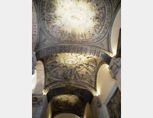Interno di una chiesa o edificio storico con soffitto a volte decorate da stucchi in rilievo, medaglioni ovali e scene figurative. Archi e cornici ornate incorniciano la volta, con illuminazione calda che mette in risalto i dettagli.