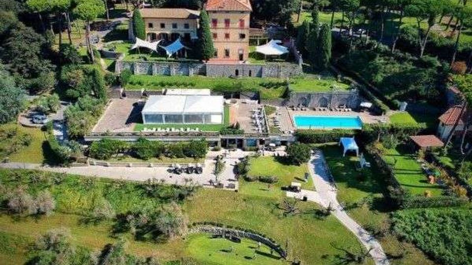 Veduta aerea di una villa storica con edifici in muratura e tetti in tegole, circondata da un ampio parco alberato. In primo piano si vedono terrazze con tensostrutture bianche, una piscina rettangolare e vialetti che attraversano prati e giardini.