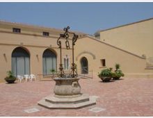 Cortile lastricato in mattoni con un pozzo in pietra al centro e struttura in ferro battuto. Sullo sfondo edifici color sabbia con grandi archi vetrati, una porta ad arco e alcune piante in vaso lungo i lati.