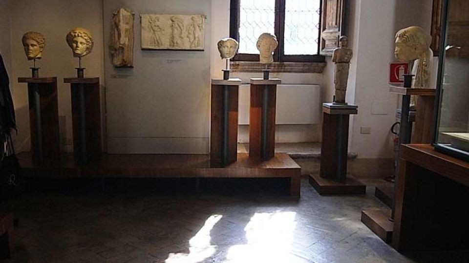 Sala museale con pareti bianche e pavimento in pietra, illuminata dalla luce che entra da una grande finestra ad arco con grate. Diverse teste scultoree su piedistalli e rilievi in pietra sono esposti lungo le pareti e su piattaforme in legno.