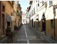 Strada stretta lastricata in un centro storico, fiancheggiata da edifici alti con persiane e balconi, con tavolini all’esterno e cavi/lanterne lungo le facciate. In fondo alla via si vede la facciata di una chiesa con portale centrale e decorazioni.