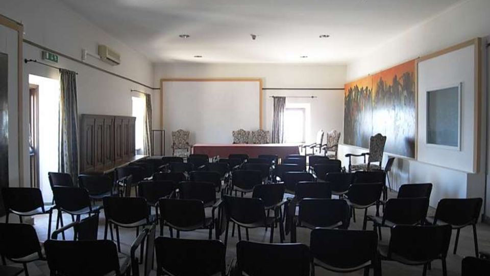 Sala riunioni con file di sedie nere rivolte verso un palco con tavolo e grande schermo di proiezione. Pareti chiare con finestre laterali e tende, armadio in legno a sinistra e grande dipinto murale sulla parete destra.