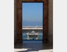 Veduta attraverso un grande portale in pietra che incornicia una terrazza monumentale con balaustra e una struttura decorativa centrale. Sullo sfondo si vede una valle con campagna e, più lontano, montagne sotto un cielo sereno.