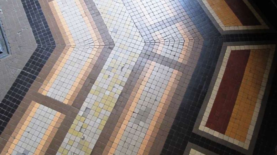 Pavimento interno in mosaico di piccole piastrelle quadrate, con motivi geometrici a bande e riquadri in tonalità grigio, beige, ocra e bordeaux. La composizione crea linee angolate e cornici che si incrociano lungo il corridoio.