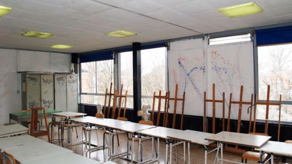 Aula scolastica vuota con file di banchi e sedie, armadietti metallici e pareti con segni e graffiti. Lungo un lato ci sono grandi finestre con infissi scuri e pannelli oscuranti blu, con cavalletti da disegno appoggiati vicino ai vetri.