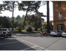 Parcheggio e incrocio stradale davanti a un’area verde con alberi alti e cespugli. Sul lato destro si vede la facciata di un edificio in intonaco arancione con finestre, e lungo la strada sono presenti auto e motociclette parcheggiate.