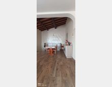 Sottotetto adibito a zona pranzo con pavimento in legno, soffitto inclinato con travi a vista e pareti chiare. Al centro c’è un tavolo con sedie; sul lato destro un mobile basso con oggetti e in fondo una ringhiera metallica che delimita l’apertura verso il piano inferiore.