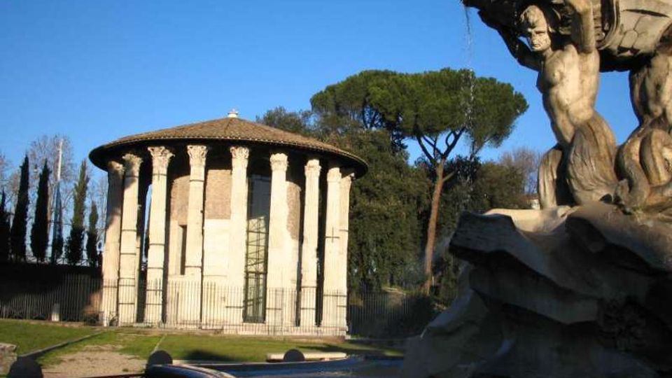 Edificio circolare in muratura con portico di colonne classiche e tetto a falde, situato in un’area verde con alberi alti sullo sfondo. In primo piano, una fontana/scultura in pietra con figure e vasca curvilinea.