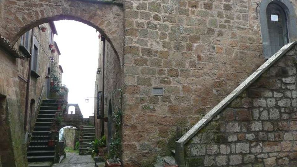 Vicolo acciottolato in un borgo storico, stretto tra edifici in pietra con archi di collegamento sopraelevati. Scale in pietra su entrambi i lati e vasi con piante lungo le pareti, con un altro arco visibile sullo sfondo.