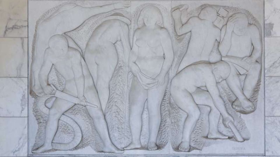 Bassorilievo in pietra chiara montato su una parete in marmo, con figure umane stilizzate disposte in più scene su un pannello rettangolare. Le sagome sono scolpite in basso rilievo con superfici lisce e sfondo leggermente incavato.