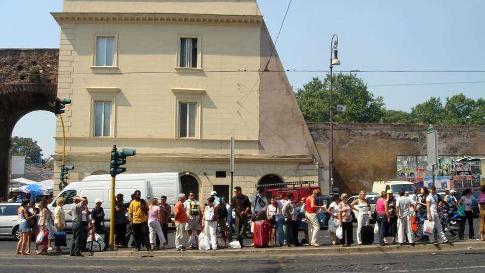 Edificio cittadino di tre piani color crema con finestre rettangolari e portico ad archi al piano terra, affacciato su una strada trafficata. Davanti si vedono marciapiede con semaforo e lampione, un arco in muratura e un muro con manifesti sullo sfondo.