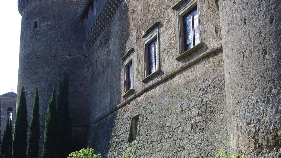 Vista dal basso di un castello in pietra con alta torre circolare e mura massicce, con finestre rettangolari e mensole sotto il tetto. In primo piano c’è un prato con cespugli, cipressi e alcuni grandi vasi in terracotta.
