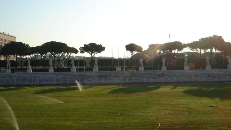 Ampio campo sportivo in erba con irrigatori in funzione e pista di atletica in primo piano. Sullo sfondo si vede una gradinata in pietra con statue allineate, alberi a chioma piatta e cielo luminoso.