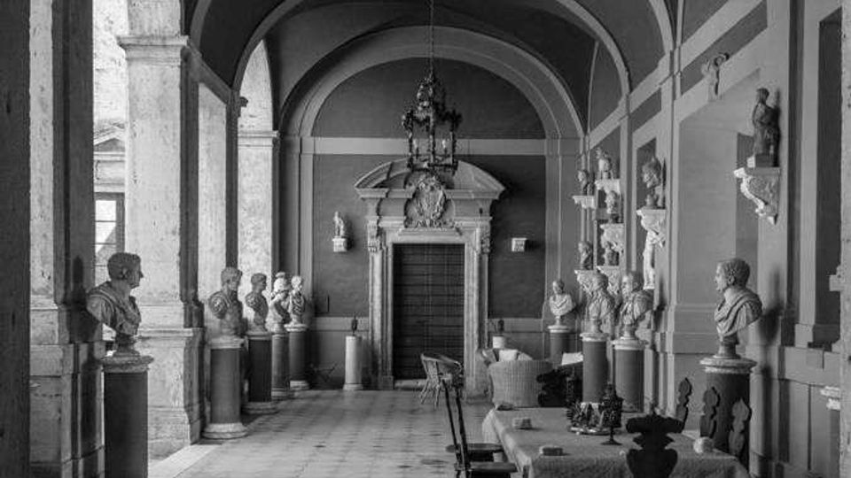 Corridoio interno con volte a botte e grandi archi in pietra, pavimento a piastrelle e una porta monumentale sul fondo. Lungo le pareti e su piedistalli sono esposti numerosi busti e statue, con tavoli e sedie disposti al centro sotto un lampadario.