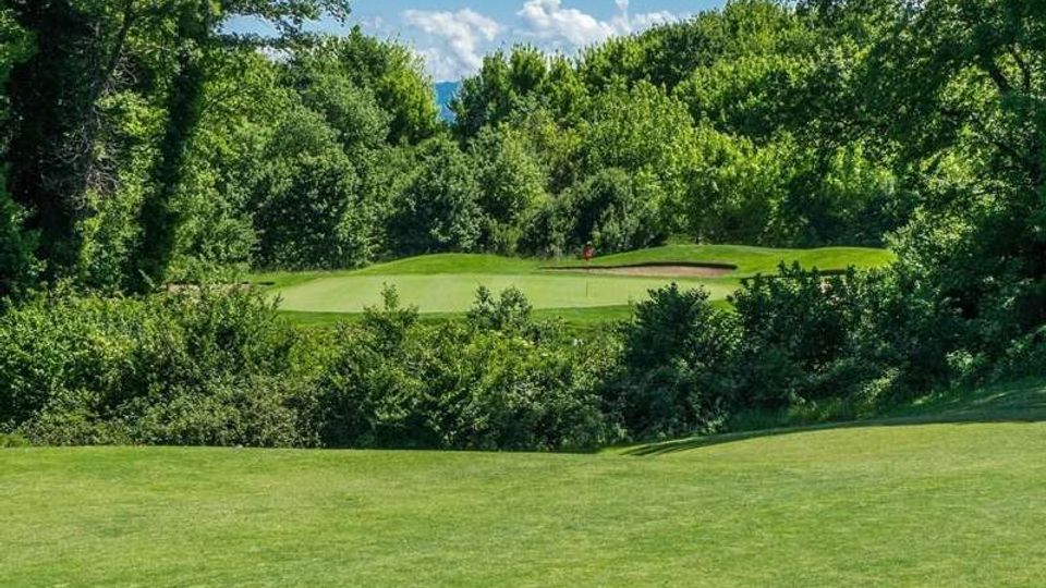 Veduta di un campo da golf con ampio prato rasato e green centrale, affiancato da un bunker di sabbia. L’area è circondata da fitta vegetazione e alberi ad alto fusto, con cielo azzurro e nuvole sullo sfondo.