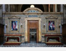 Interno di una chiesa con parete in marmi policromi e portale centrale ad arco che conduce a una cappella. Ai lati del portale ci sono due grandi dipinti incorniciati, lampade a parete e nicchie con sarcofagi, sopra un’iscrizione decorata con stelle e una piccola finestra circolare.