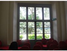 Sala interna con file di sedie imbottite rosse (e una nera) rivolte verso una grande porta-finestra a più riquadri. Attraverso i vetri si vede un’area verde con alberi e vegetazione, con segnaletica di uscita sulla parete a destra.