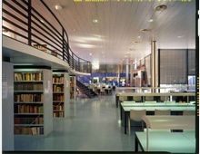 Interno di una biblioteca moderna con scaffali di libri, tavoli di studio con lampade e sedie disposti in file. Si vedono un soppalco con ringhiera metallica e una scala che conduce al livello superiore, sotto un soffitto alto con illuminazione integrata.