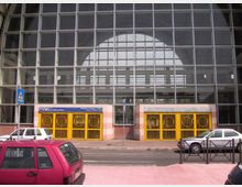 Facciata di un grande edificio pubblico con ampia vetrata a griglia che copre un alto arco centrale. Alla base ci sono due ingressi con cancelli gialli, insegne sopra le porte e marciapiede su strada con auto parcheggiate e segnaletica pedonale.