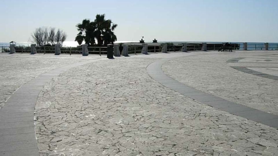 Ampia piazza panoramica pavimentata in pietra chiara con percorsi curvilinei in lastre scure, affacciata sul mare tramite una ringhiera. Sullo sfondo si vedono una palma, alberi spogli e alcune panchine lungo il belvedere.