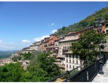 Vista panoramica di un borgo collinare con edifici in pietra e intonaco dai colori caldi disposti in terrazze lungo il pendio. In primo piano si vede una passeggiata con ringhiera e lampioni, circondata da alberi e vegetazione, con valle sullo sfondo sotto un cielo sereno.